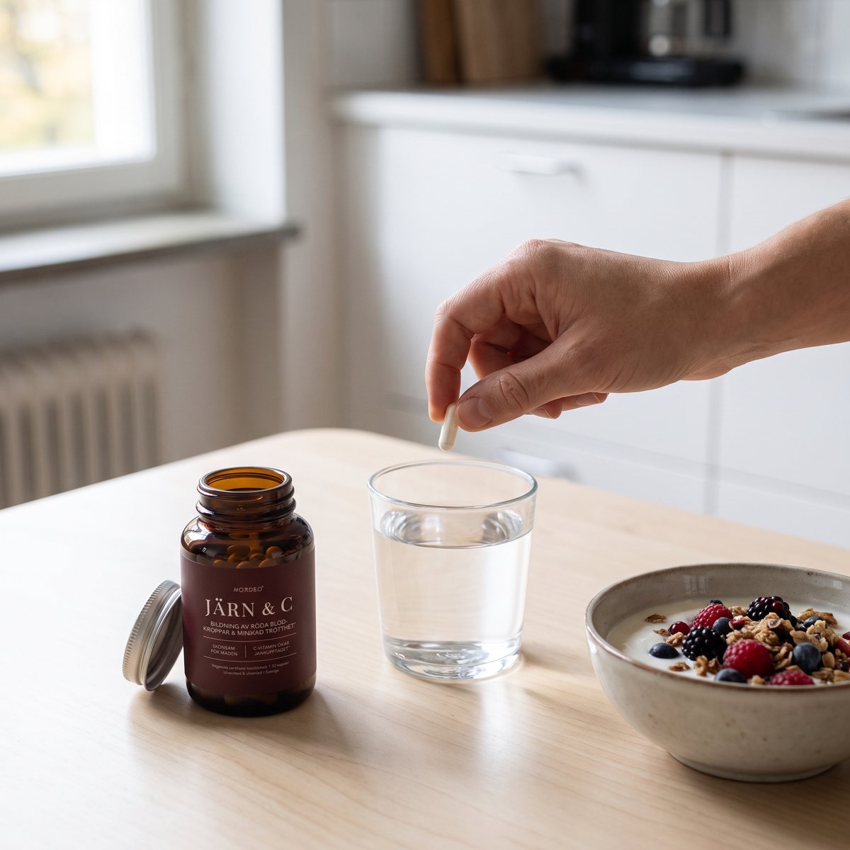Nordbo Järn & C-vitamin i mörk glasburk på ett köksbord i ljust trä bredvid ett glas vatten och en skål yoghurt med bär, en hand håller en kapsel