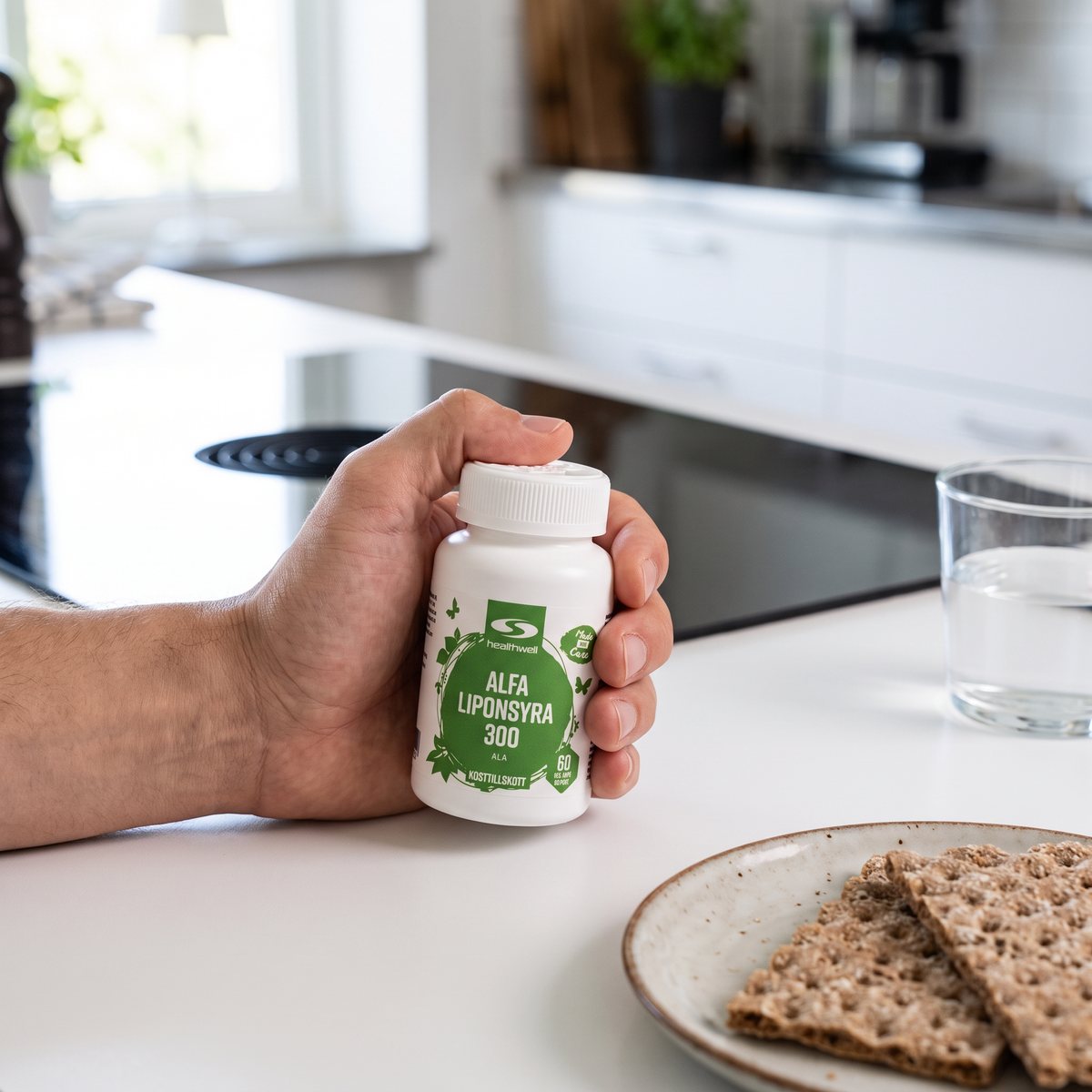 Hand som håller Healthwell Alfa Liponsyra 300 vid köksbänken med knäckebröd och vatten i ett svenskt kök