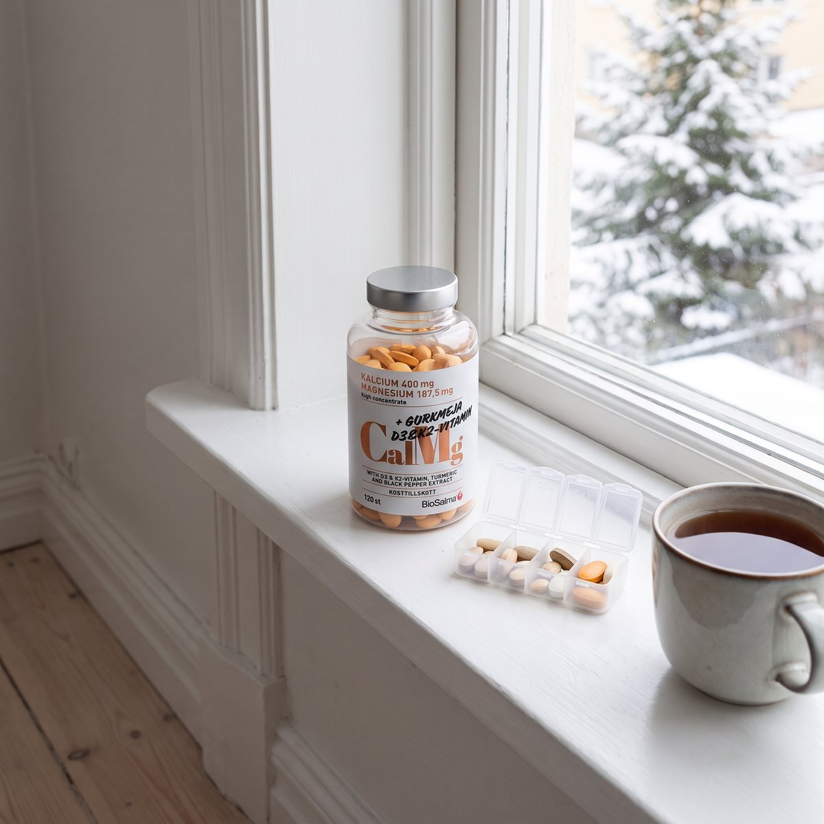 Burk med BioSalma Kalcium & Magnesium på ett vitt fönsterbräde i en svensk lägenhet, med snöig gran utanför