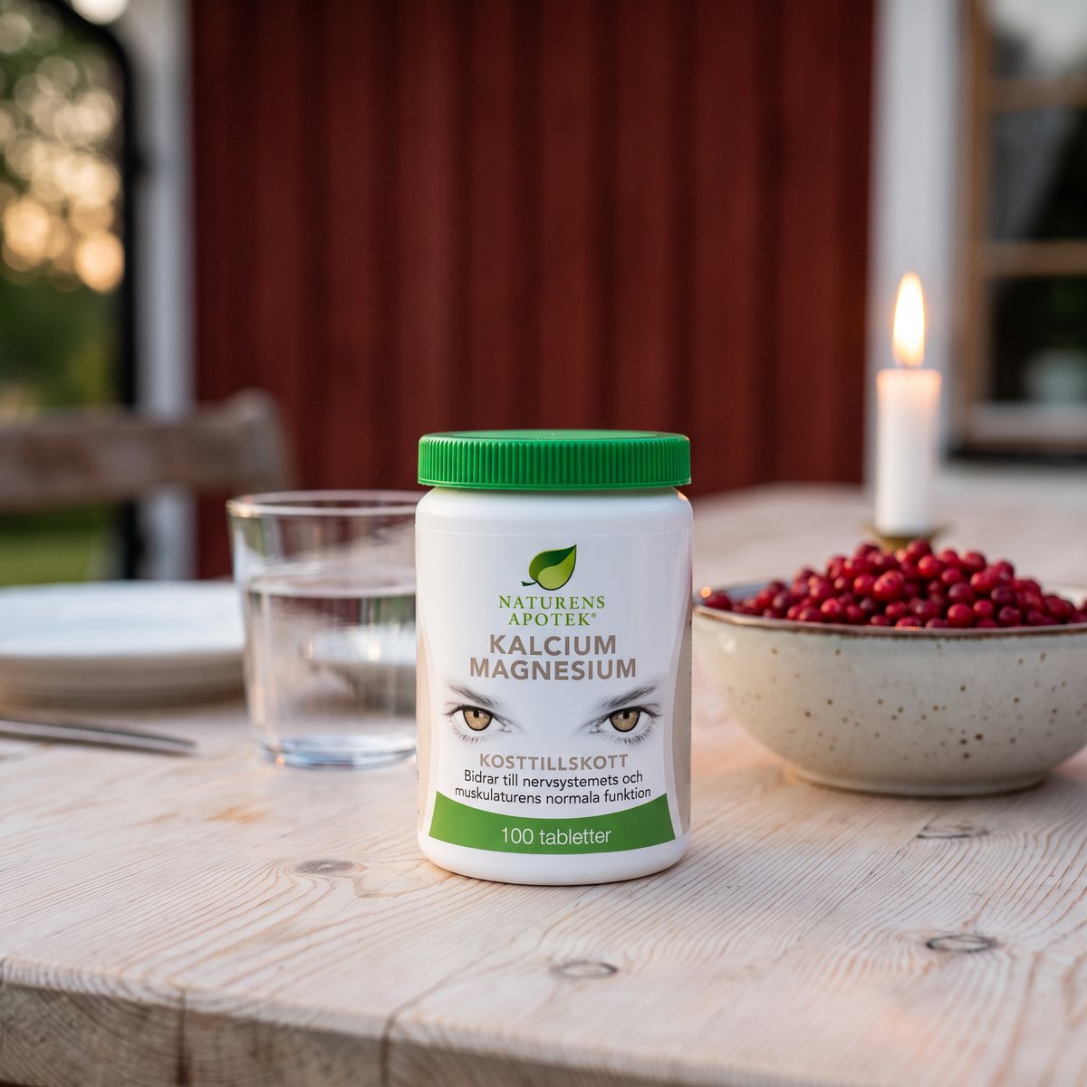Burk med Naturens Apotek Kalcium Magnesium på ett furubord vid en rödmålad stuga, med lingon och ett glas vatten