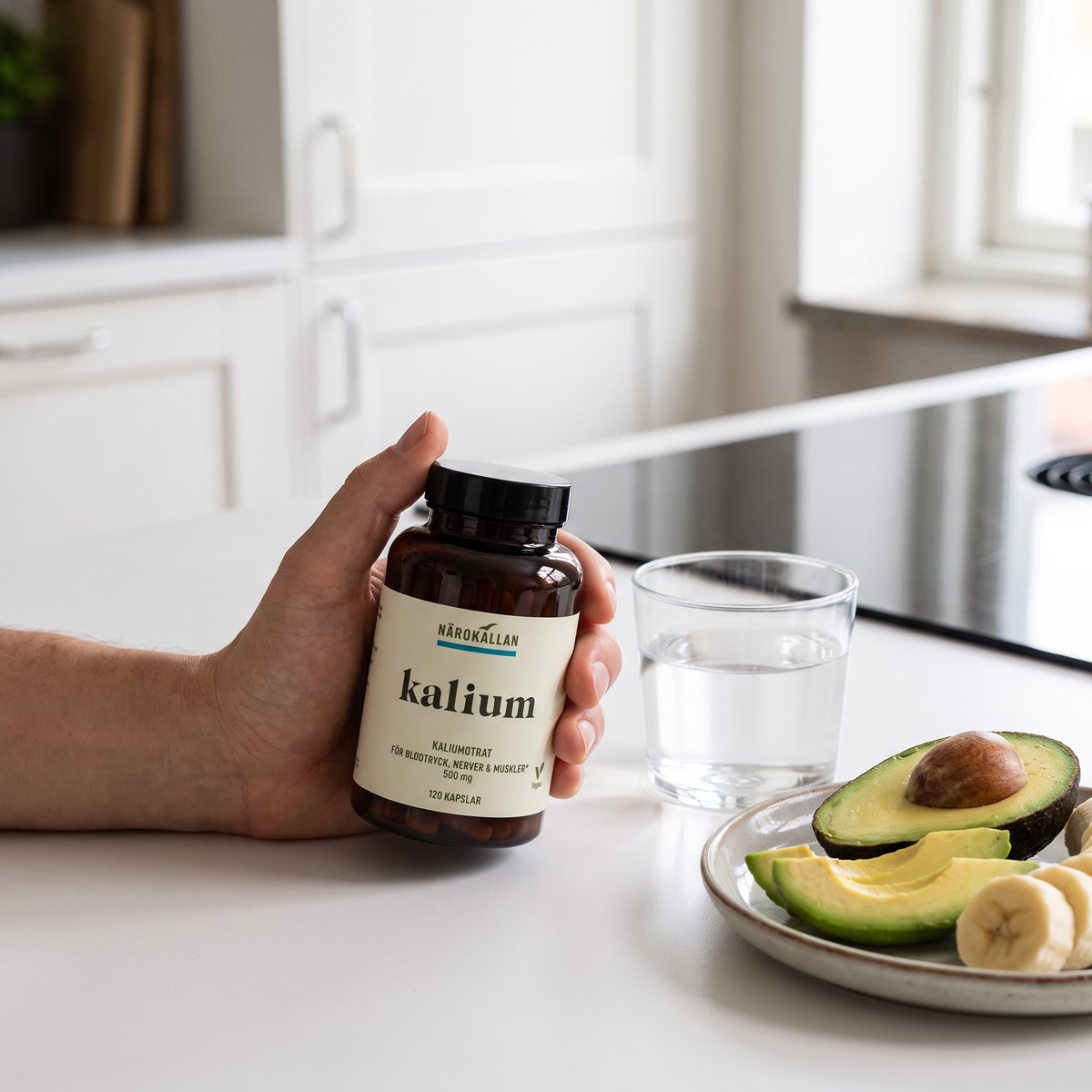 Närokällan Kaliumcitrat i brun glasburk hålls i handen vid en vit bänk i ett svenskt kök med avokado och banan