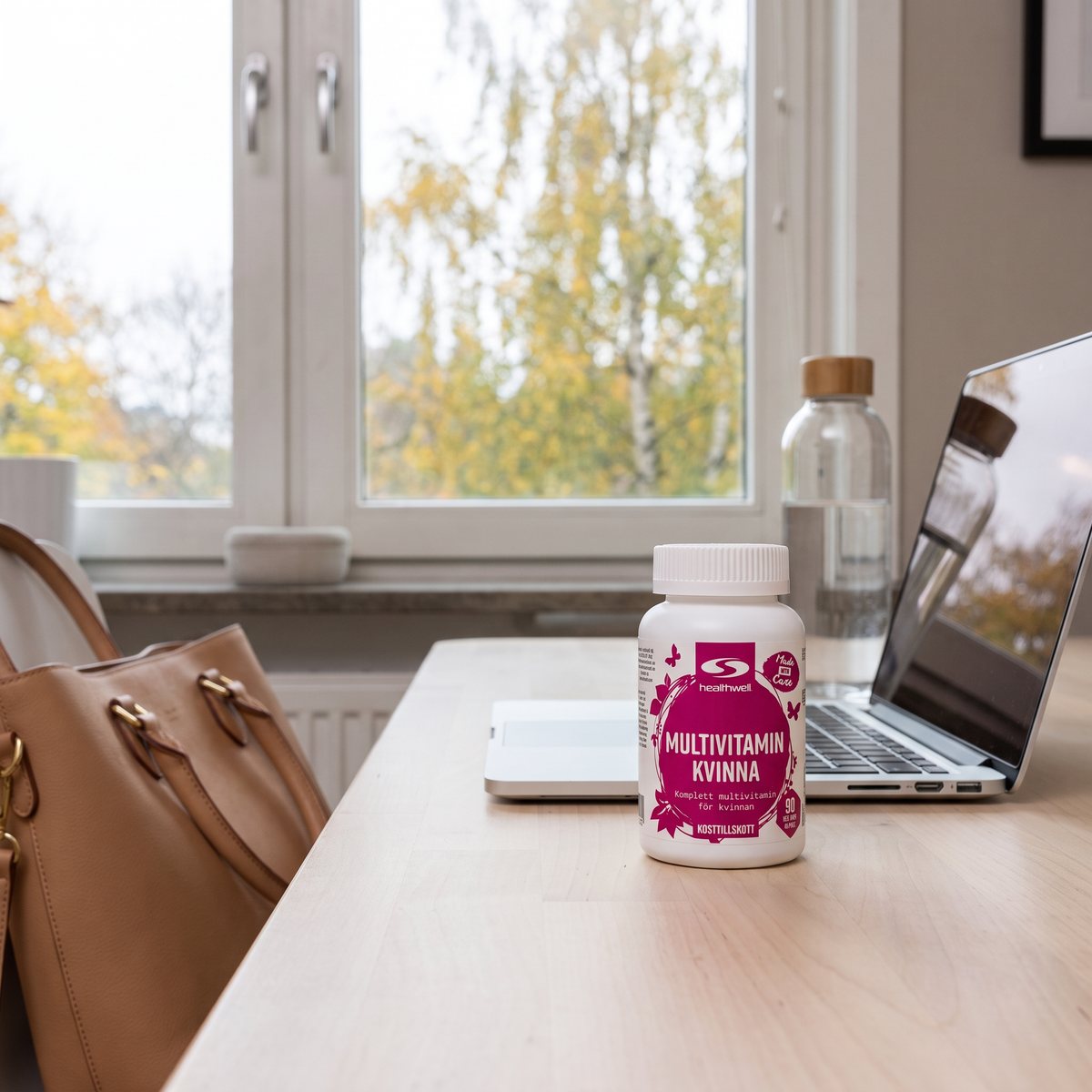 Burk Healthwell Multivitamin Kvinna på ett björkskrivbord vid laptop och vattenflaska i ett svenskt hemmakontor med höstlöv utanför