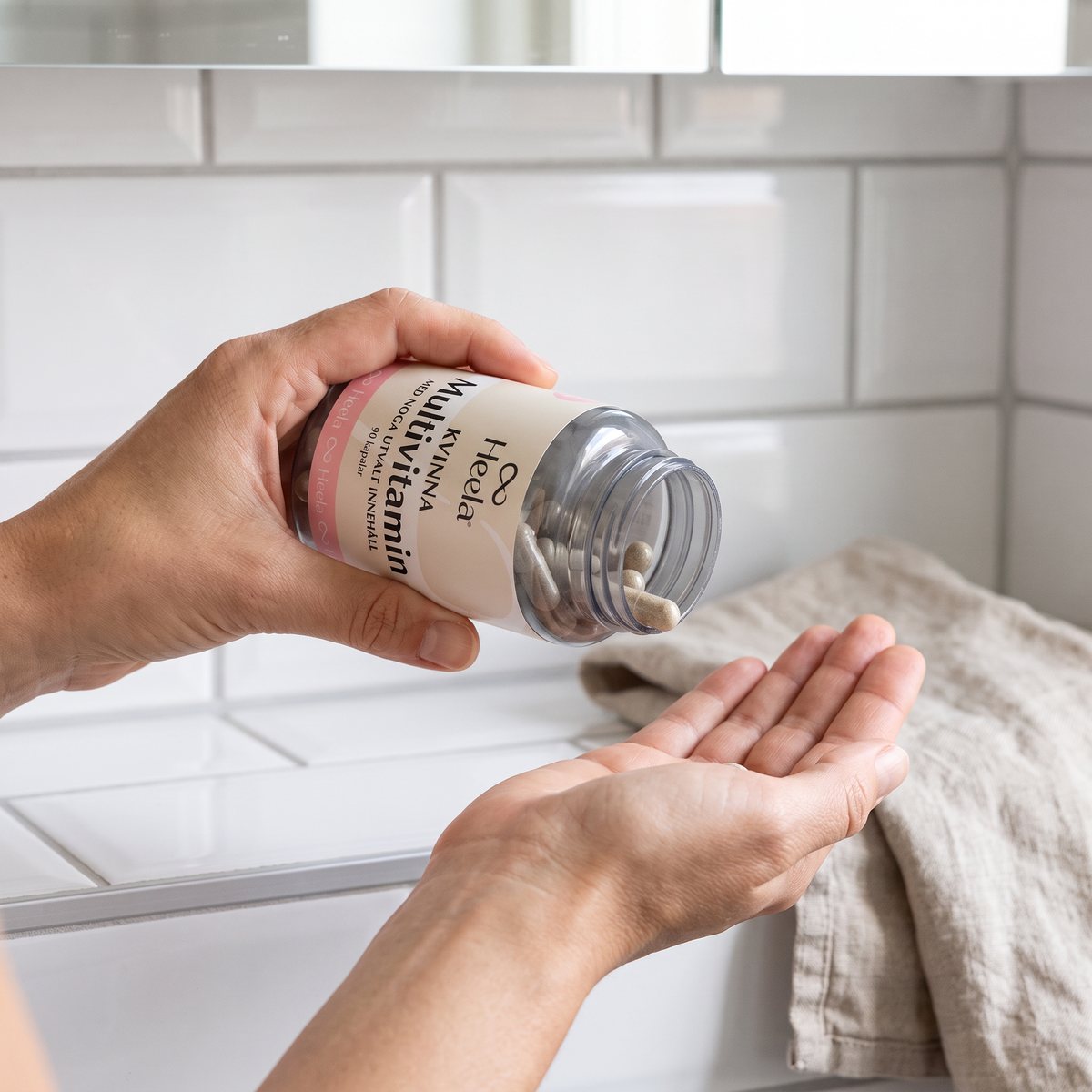Hand som skakar ut en kapsel ur Heelas multivitaminburk över handflatan, vid en vit kakelvägg i ett svenskt badrum