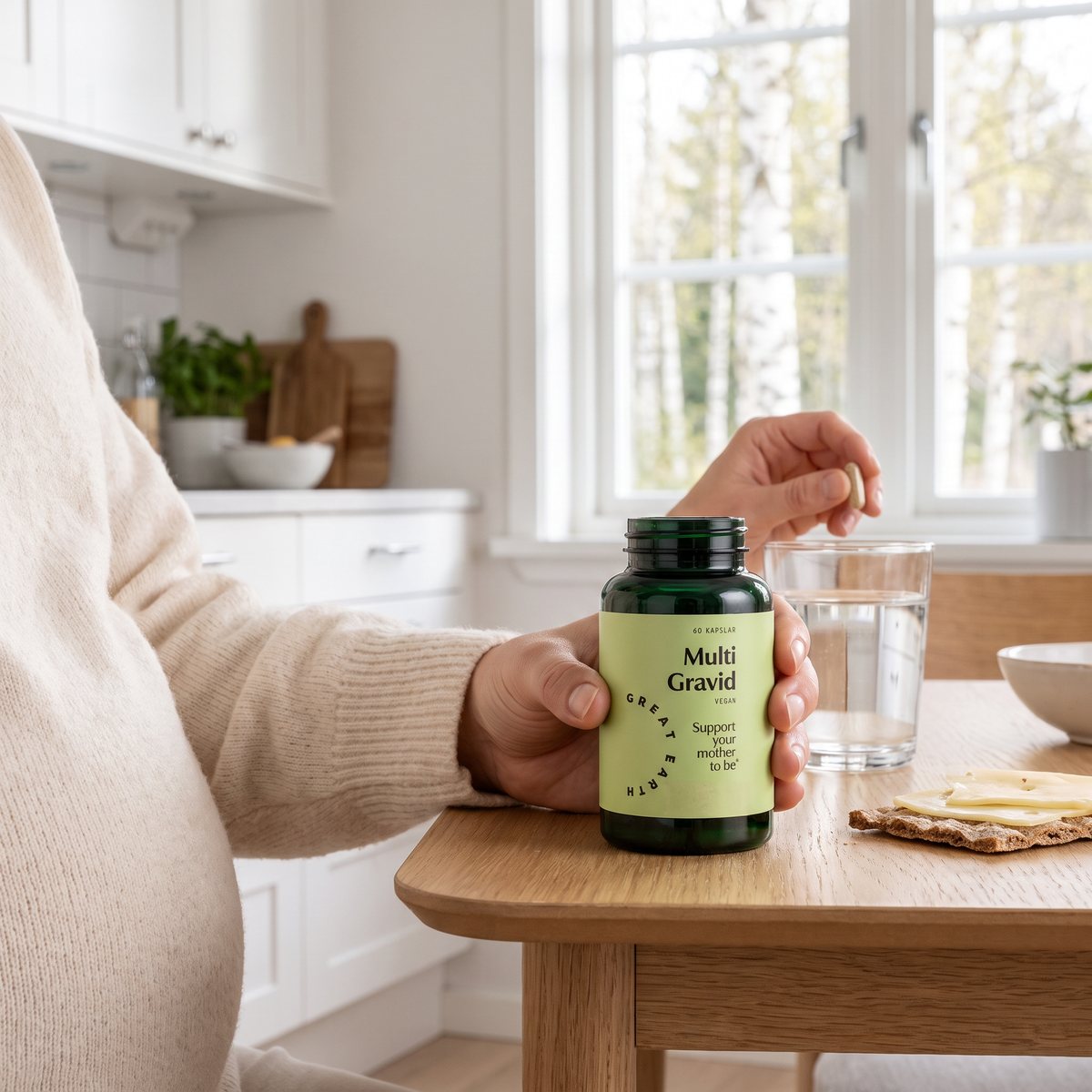 Gravid kvinna vid ett ljust köksbord håller Great Earth Multi Gravid-burken och en kapsel bredvid ett glas vatten och knäckebröd, björkar utanför fönstret