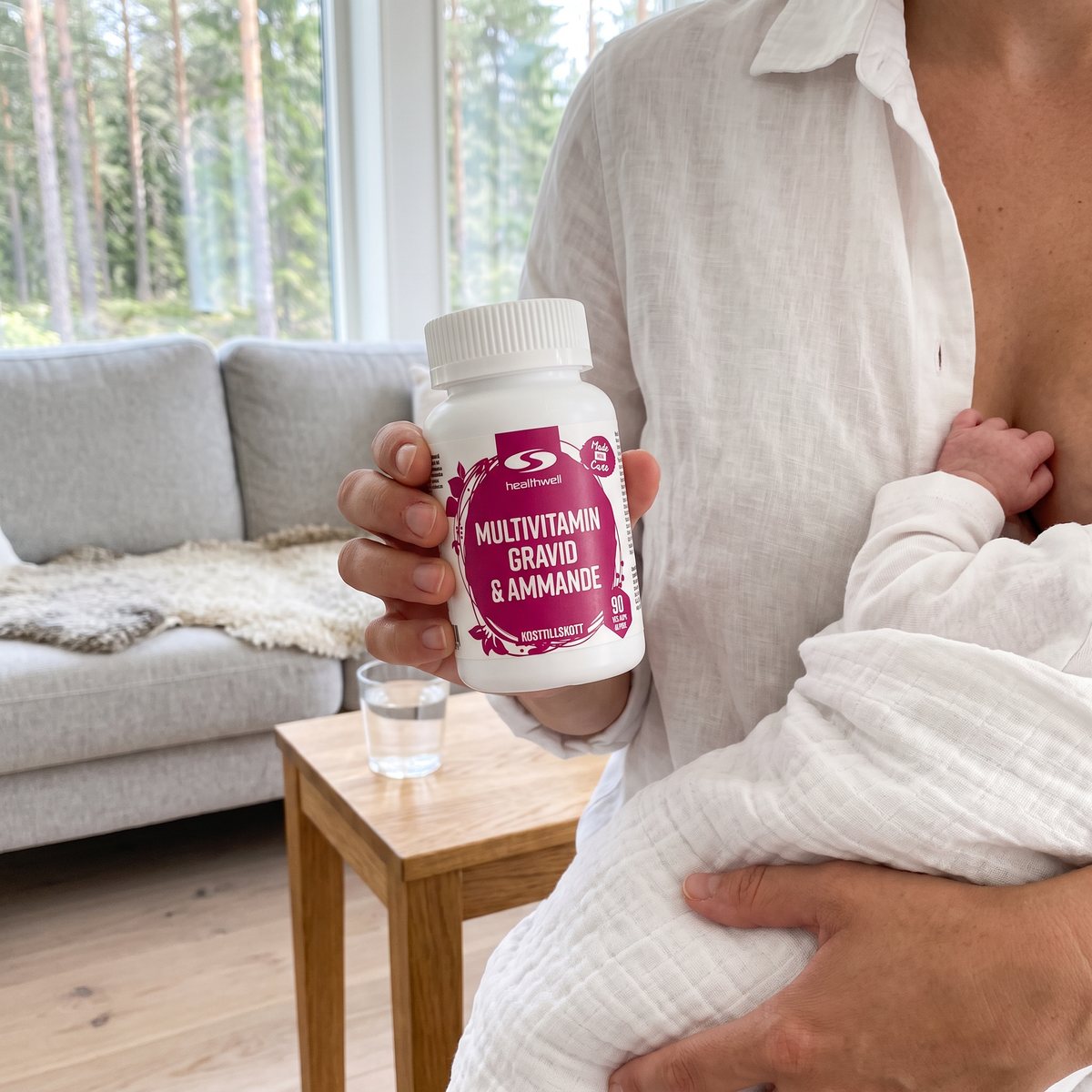 Mamma i vit linneskjorta håller ett nyfött barn i en muslinfilt och burken Healthwell Multivitamin Gravid & Ammande i ett ljust vardagsrum med tallskog utanför