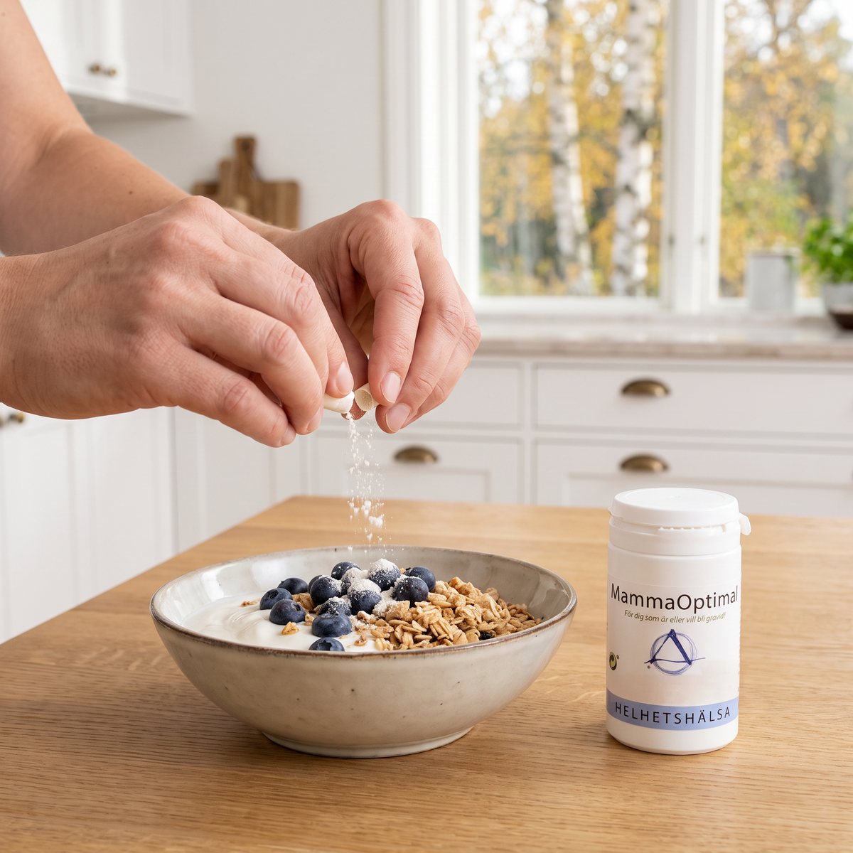 Händer drar isär en kapsel och strör pulvret över yoghurt med granola och blåbär, bredvid burken Helhetshälsa MammaOptimal på ett ekbord
