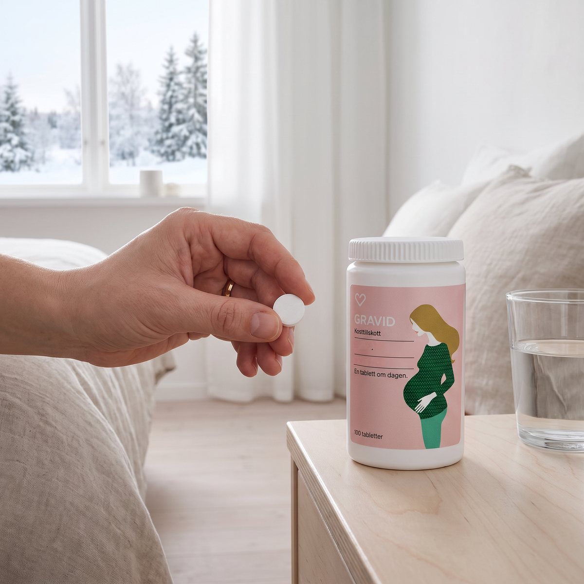 Hand håller en vit tablett bredvid burken Hjärtats Multivitamin Gravid på ett sängbord i ett ljust sovrum med snöig granskog utanför