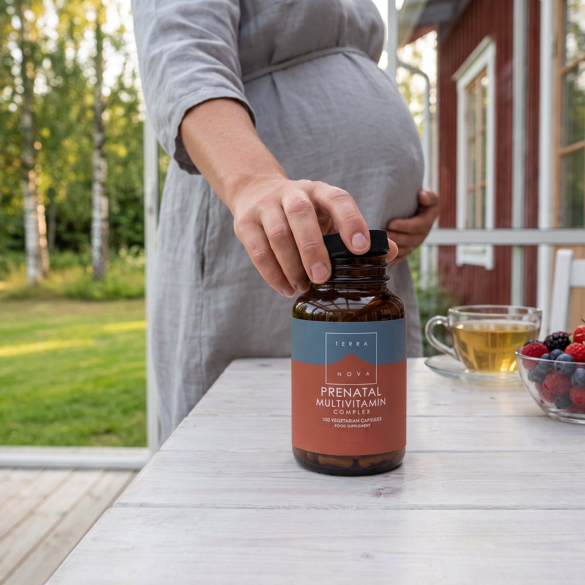 Gravid kvinna i linneklänning skruvar av locket på Terranova Prenatal Multivitamin Complex på en vit veranda vid en röd stuga med björkar