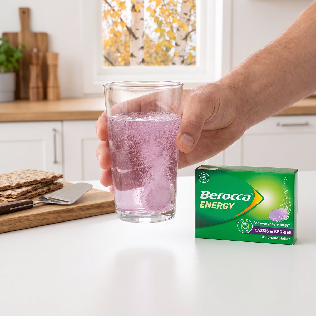 Hand som håller ett glas med lila brusdryck bredvid en grön förpackning Berocca Energy Cassis & Berries i ett ljust svenskt kök med knäckebröd och osthyvel