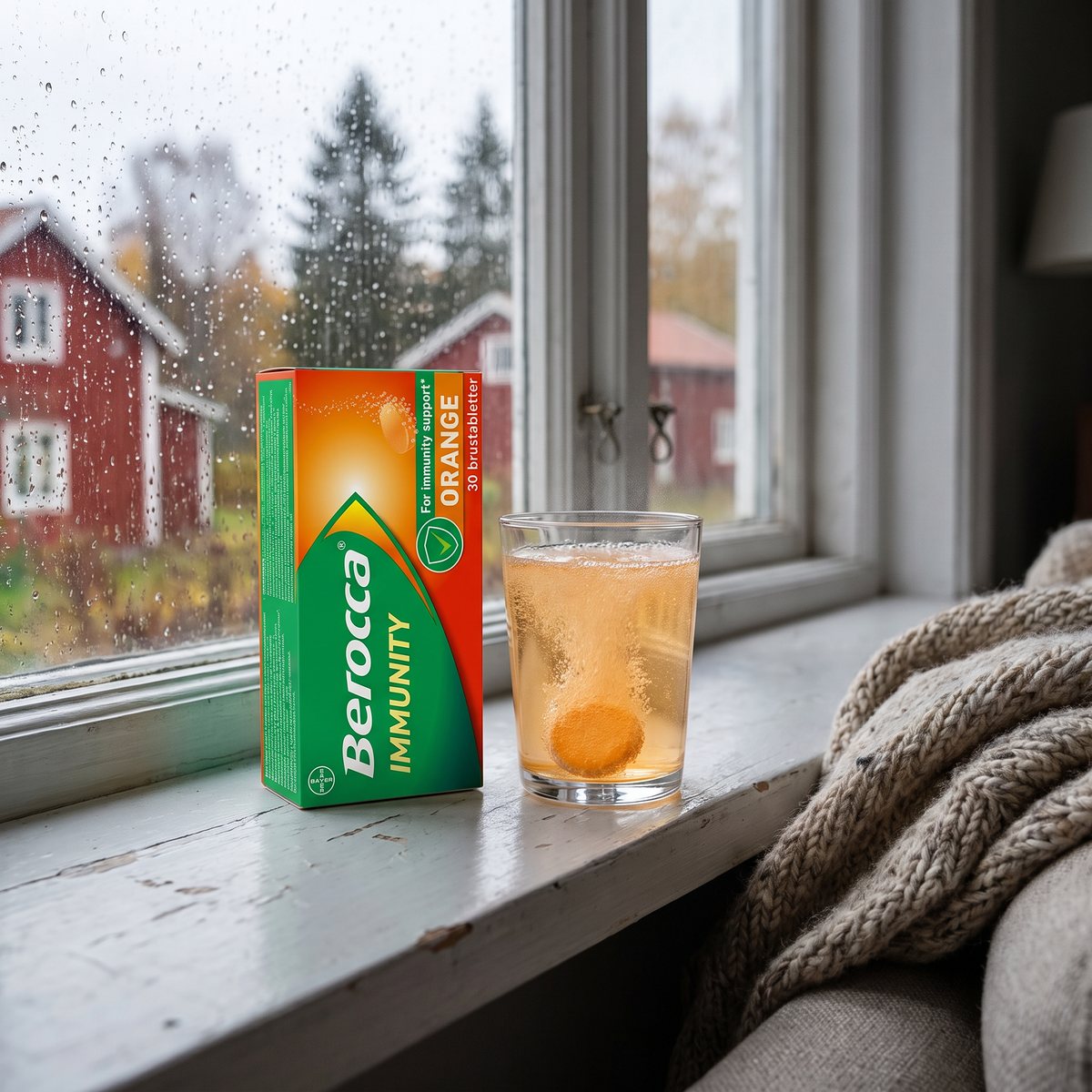Förpackning Berocca Immunity Orange och ett glas med apelsinfärgad brusdryck på en fönsterbräda, med röda trähus och granar i regnet utanför