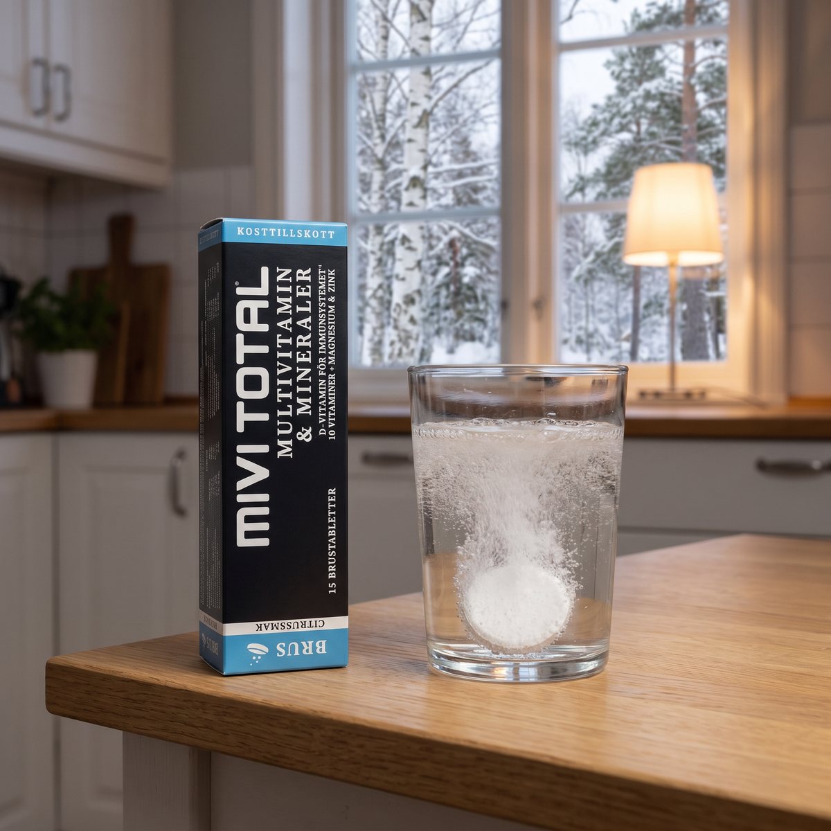 Svart förpackning med Mivitotal brustabletter bredvid ett glas vatten där en tablett löses upp, på en ekbänk i ett svenskt kök en snöig vintermorgon