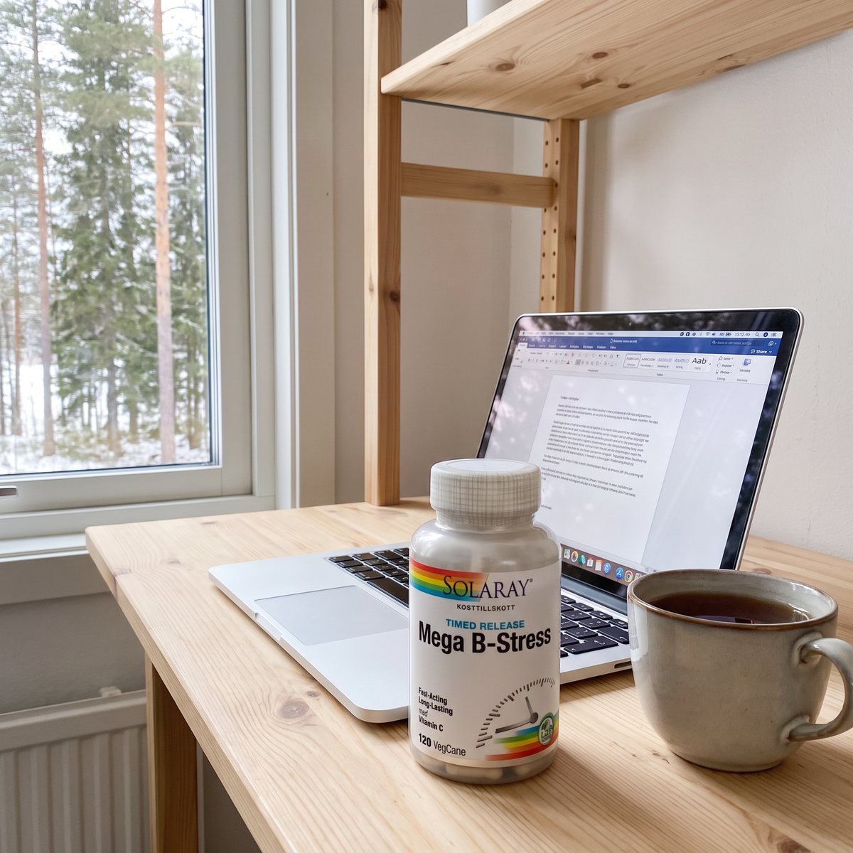 Burk med Solaray Mega B-Stress på ett skrivbord framför en laptop och en tekopp, med tallskog utanför fönstret i ett svenskt hem