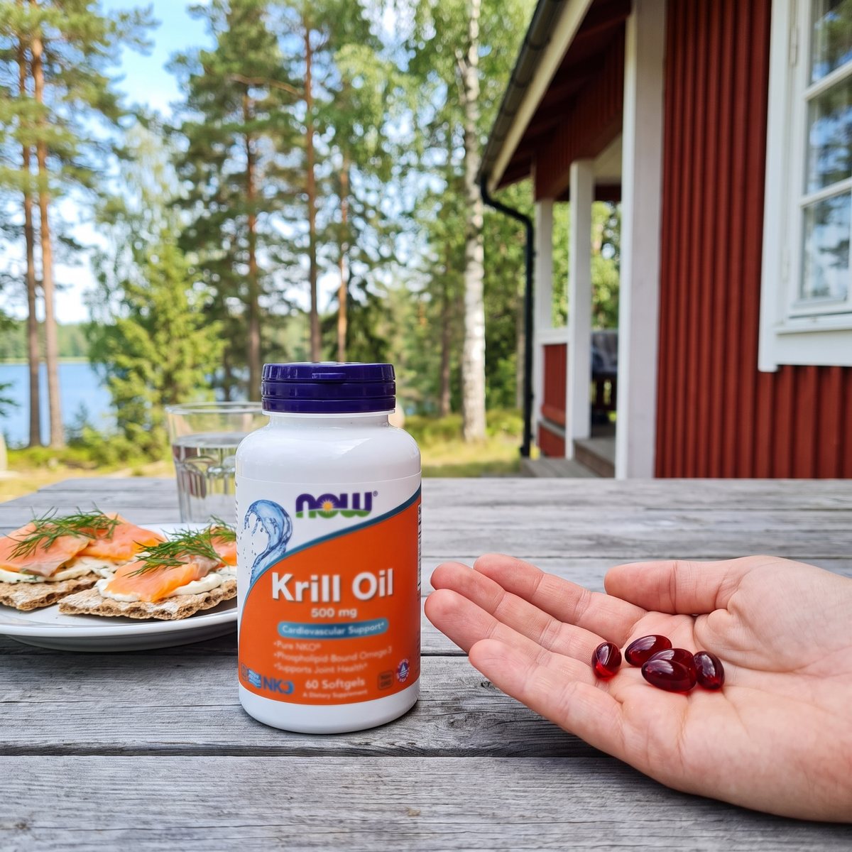 Burk NOW Krillolja 500 mg på ett grått träbord vid en röd sommarstuga med tallar och sjö, bredvid knäckebröd och en hand med några kapslar