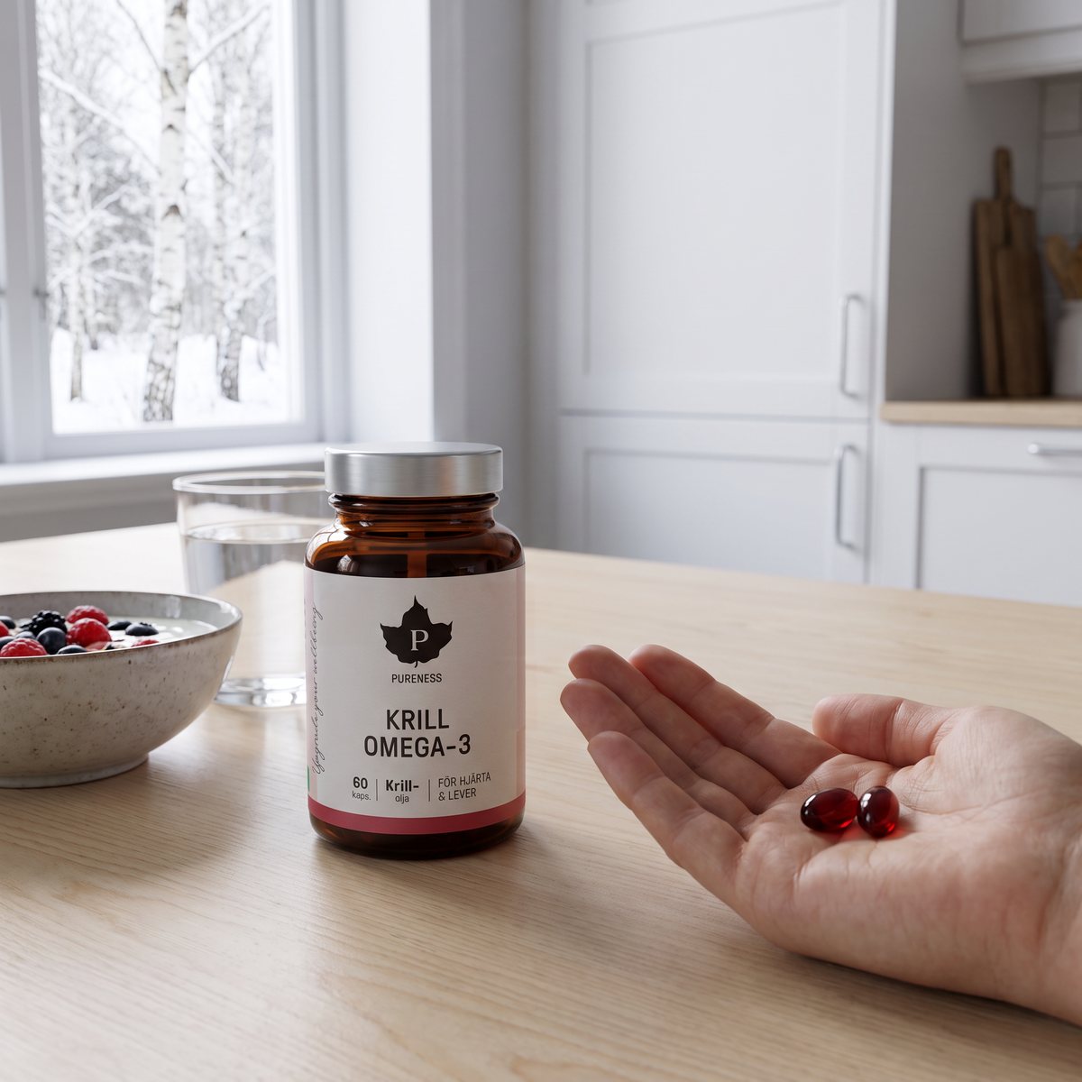 Burk med Pureness Krill Omega-3 på ett köksbord i ljus björk bredvid yoghurt och ett glas vatten, en hand håller två krilloljekapslar och snöiga björkar syns genom fönstret