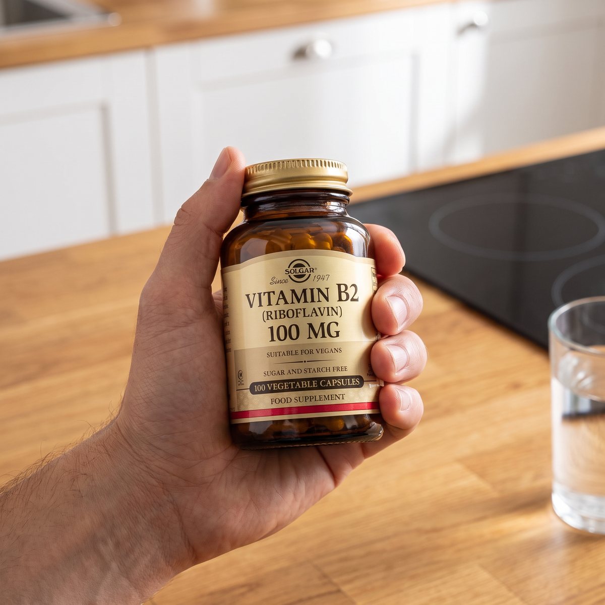 Hand som håller en brun glasburk med Solgar Vitamin B2 över en träbänk i ett svenskt kök med induktionshäll
