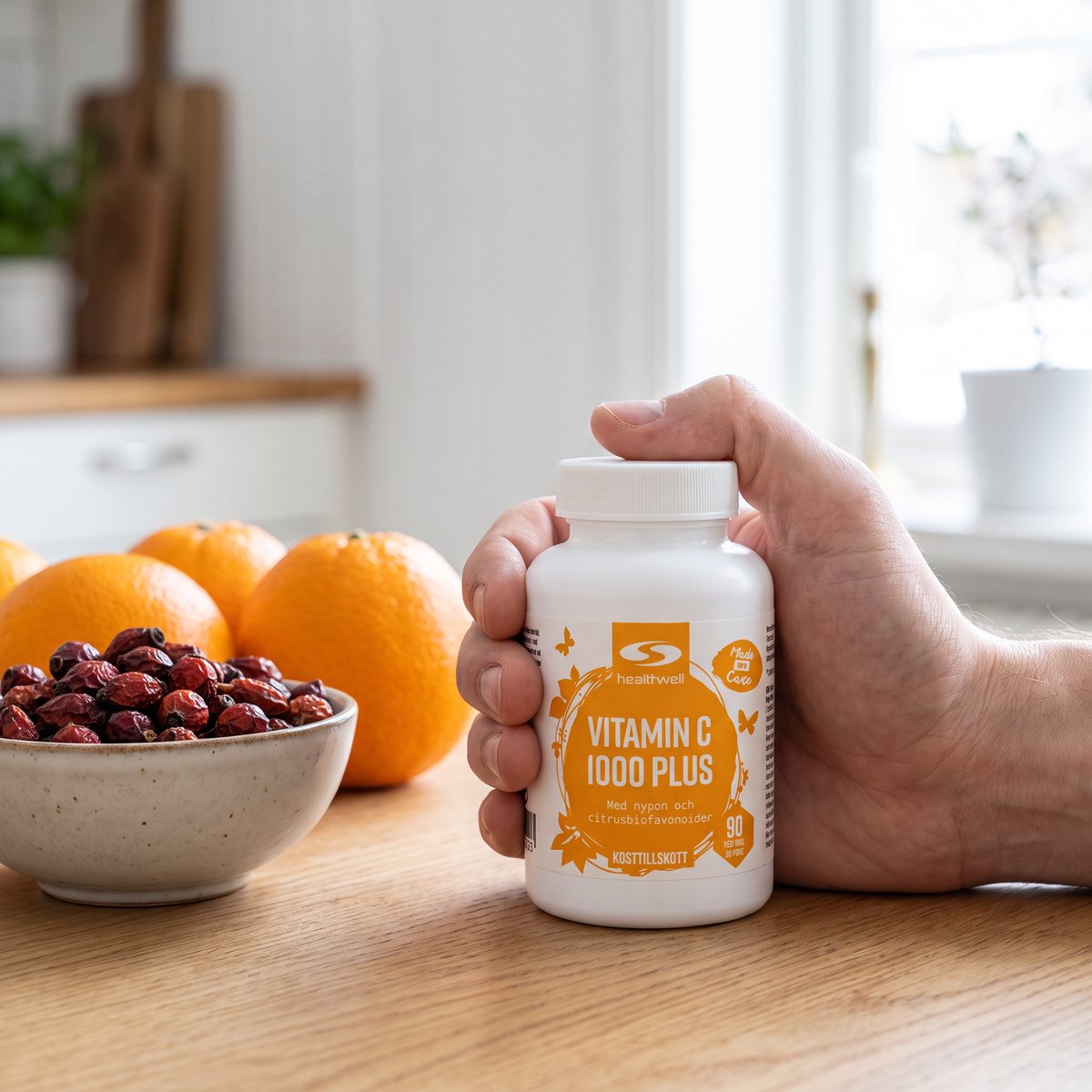 Hand som håller en burk Healthwell Vitamin C 1000 Plus vid en skål nypon och apelsiner i ett ljust svenskt kök