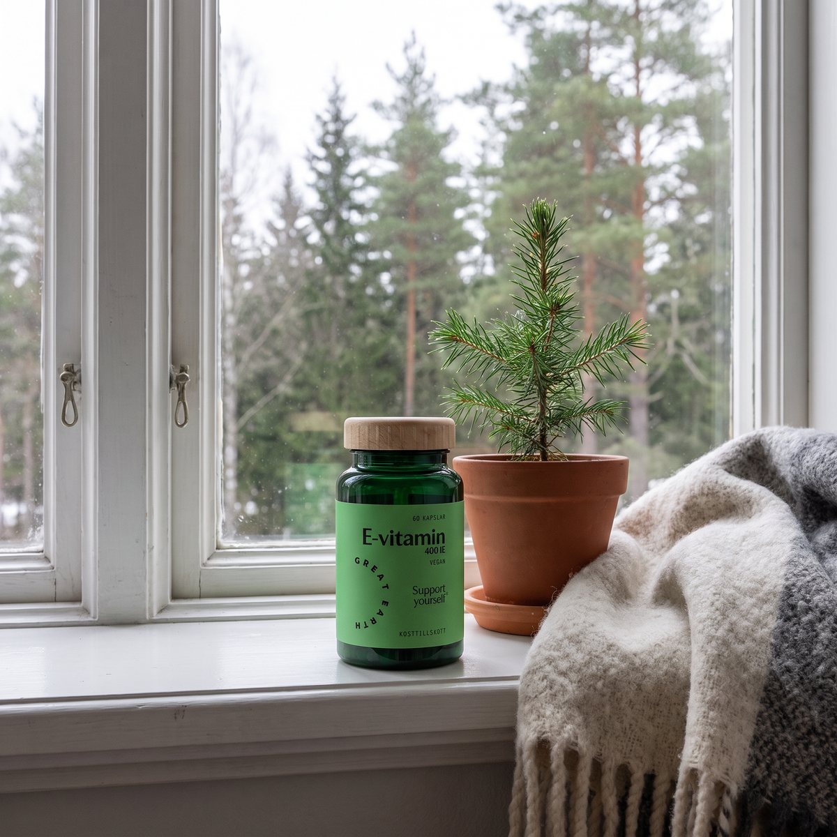 Grön glasflaska med Great Earth E-vitamin på ett fönsterbräde i en svensk lägenhet med tallar utanför