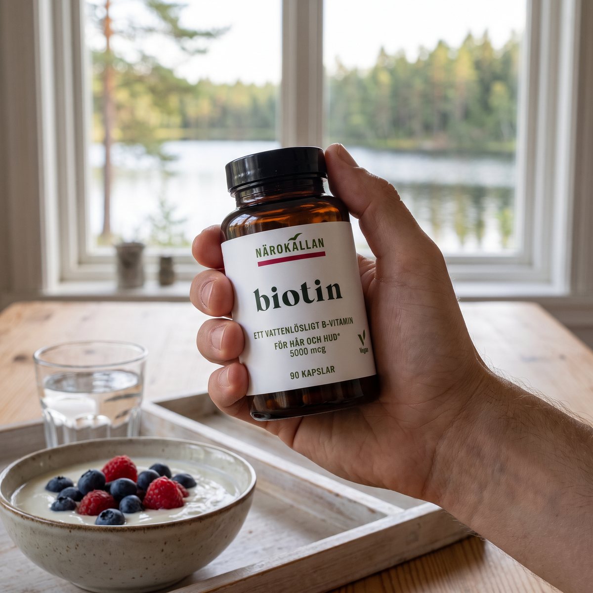 Hand som håller Närokällan Biotin-burken vid ett frukostbord med utsikt över svensk sjö och tallskog