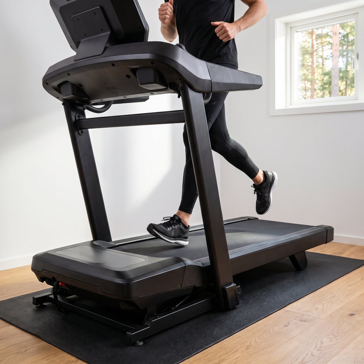 Svart Bowflex T9-löpband med löpare i ett ljust källargym i en svensk villa med trägolv och tallar utanför fönstret