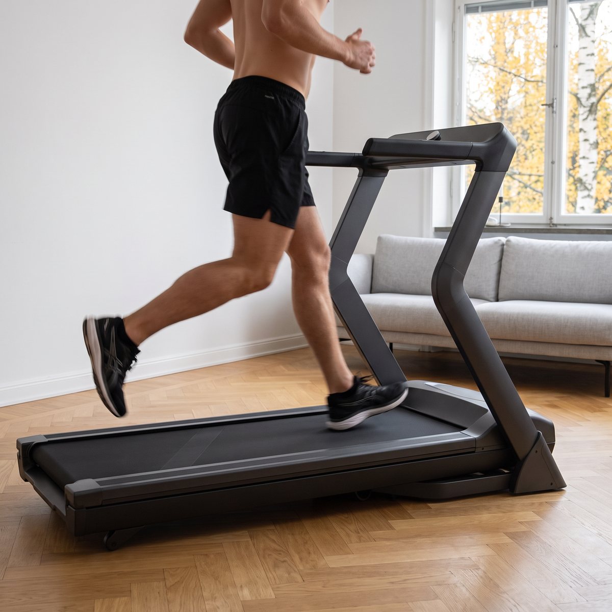 Grafitgrått Master Fitness T26 Nero-löpband med löpare i ett svenskt vardagsrum med ekparkett och björkar utanför fönstret