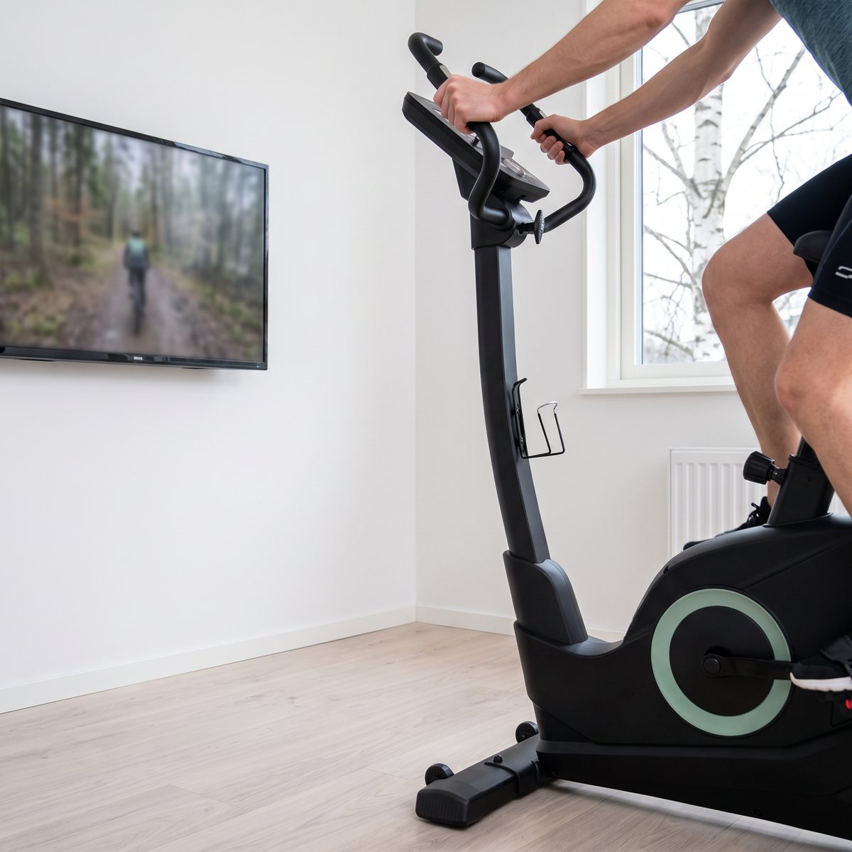 Titan Life B56 motionscykel framför en väggmonterad tv i en svensk lägenhet med björk utanför fönstret