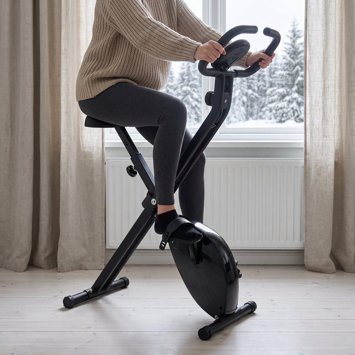 Person trampar lugnt på en svart Gorilla Sports X Frame i ett ljust sovrum med linnegardiner och snöig granskog utanför fönstret