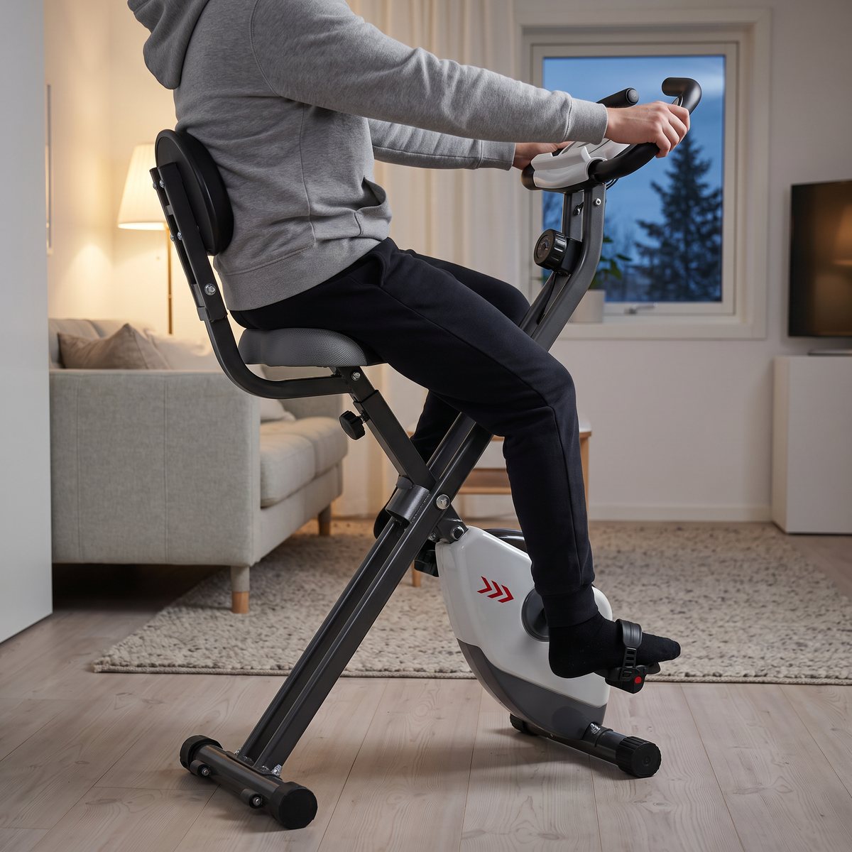 Person lutar sig mot ryggstödet och trampar på en hopfällbar Toorx BRX Compact i ett svenskt vardagsrum en vinterkväll