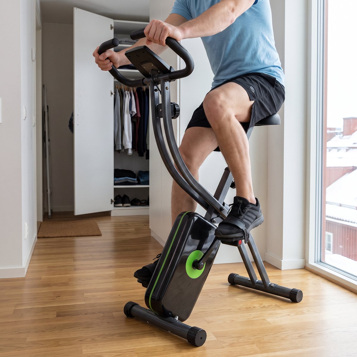 Person trampar på en hopfällbar Tunturi Cardio Fit B20 i en ljus etta med ekgolv och öppen garderob, snöiga tak utanför fönstret