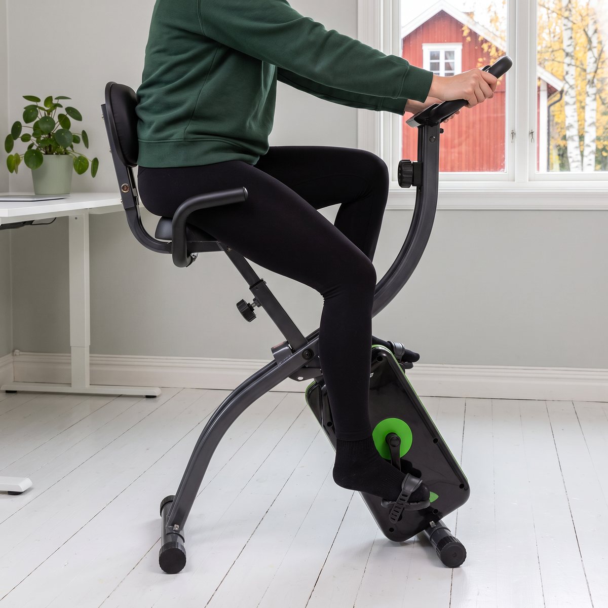 Person sitter mot ryggstödet och trampar på en Tunturi Cardio Fit B25 i ett ljust hemmakontor med vitmålat trägolv och rött trähus utanför fönstret