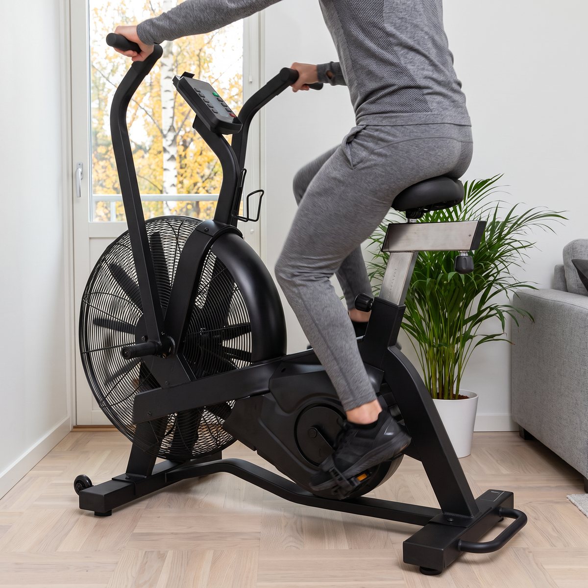 Master Fitness XB505 luftcykel i användning i ett hörn av ett ljust svenskt vardagsrum med fiskbensparkett och björkar i höstfärger utanför