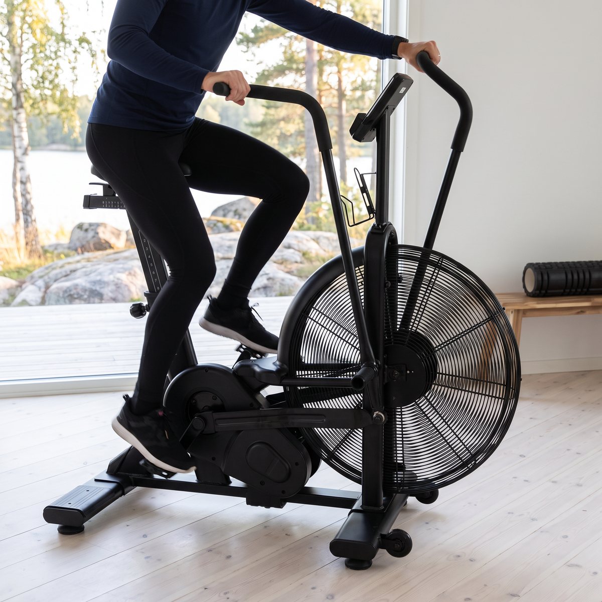 Recoil Airbike X2 luftcykel i användning i ett ljust träningsrum med vitlaserat trägolv och utsikt mot en svensk sjöstrand med granithällar och björkar