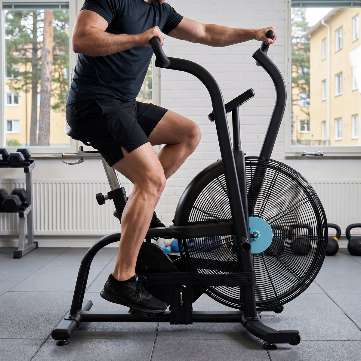 Spirit AB900+ luftcykel i användning i ett gym i en svensk bostadsrättsförening med gummigolv, hantlar och utsikt mot en innergård med tallar
