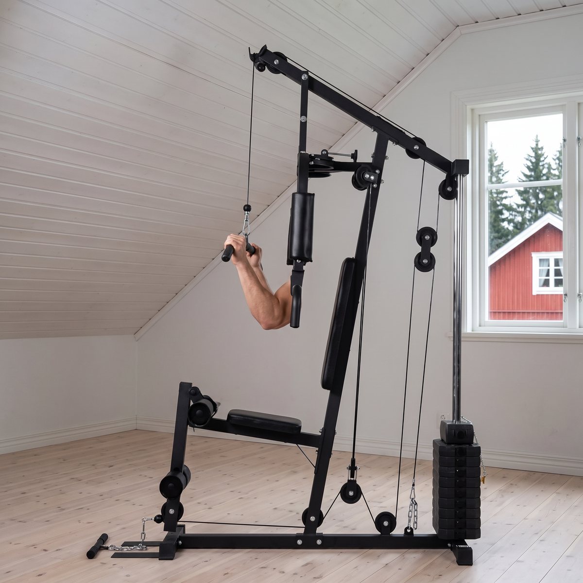 FitNord Home Gym 100 i ett svenskt vindsrum med snedtak och rött trähus utanför fönstret, använd för latsdrag
