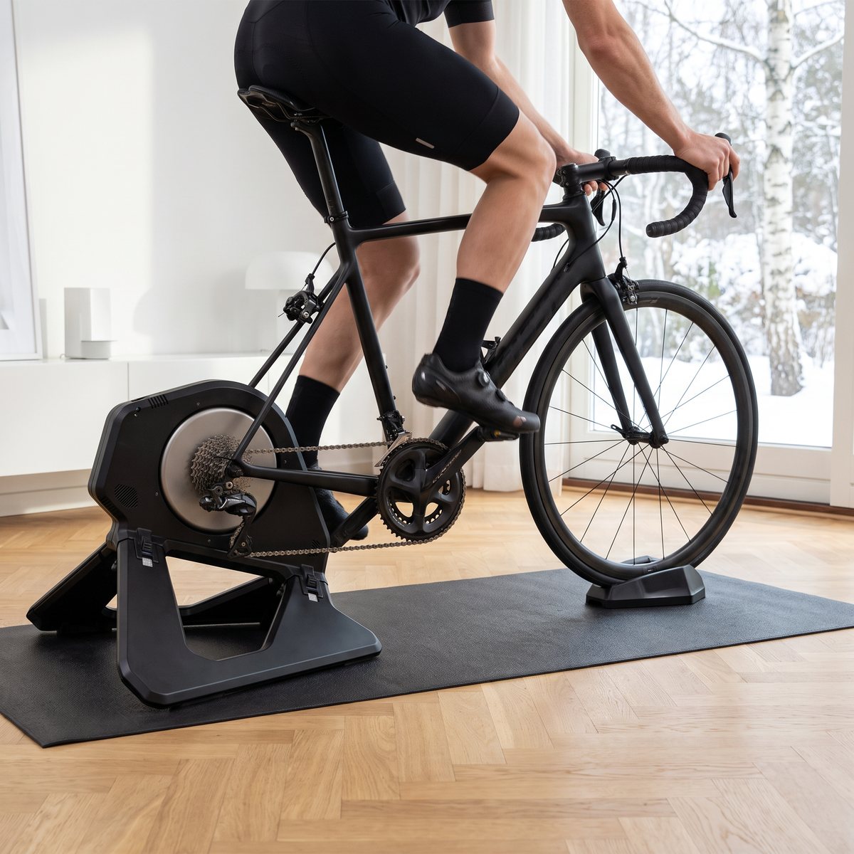 Garmin Tacx NEO 3M med en svart racercykel monterad, cyklist som trampar i ett ljust vardagsrum med fiskbensparkett och snöig björkskog utanför fönstret