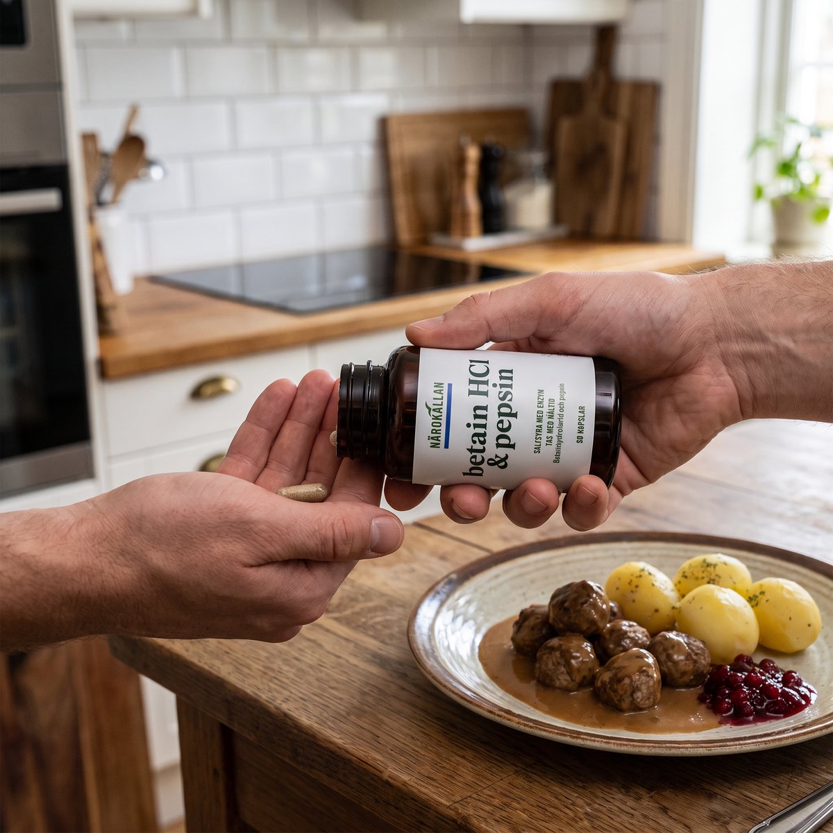 Hand som häller ut en kapsel ur Närokällans Betain HCL & Pepsin vid en tallrik med köttbullar i ett svenskt kök