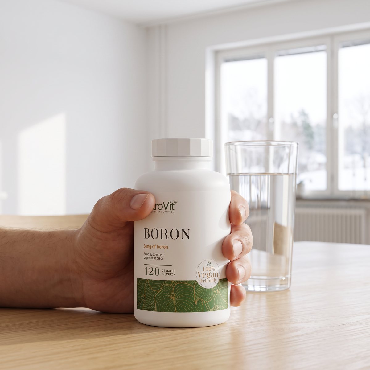 Hand som håller en burk OstroVit Boric Acid VEGE vid ett vattenglas på ett träbord i en ljus svensk bostad