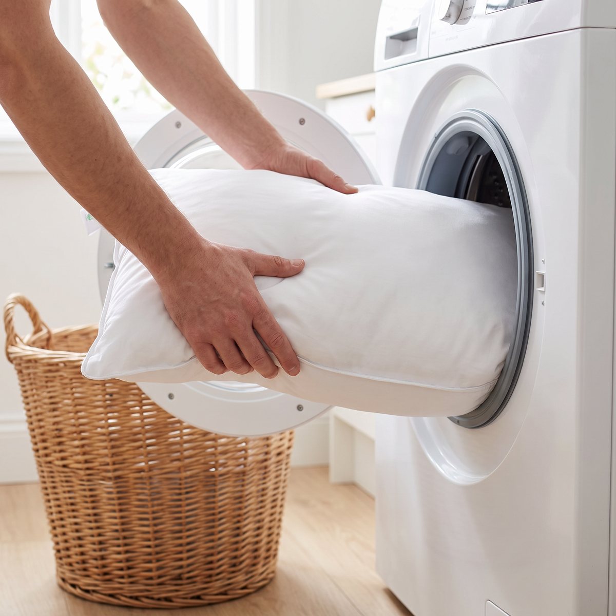 Två händer lägger en vit Borås Cotton-fiberkudde i en frontmatad tvättmaskin bredvid en tvättkorg