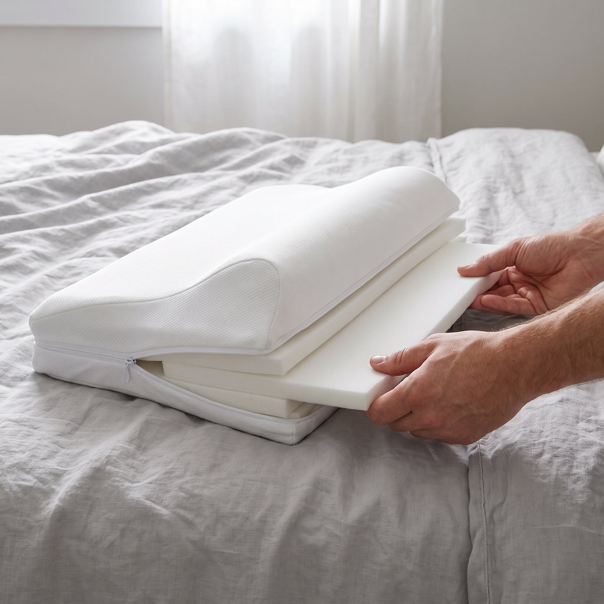 Händer som drar ut en tunn skumskiva ur en vit Comfortex NeckRest-kudde med öppen dragkedja på en säng