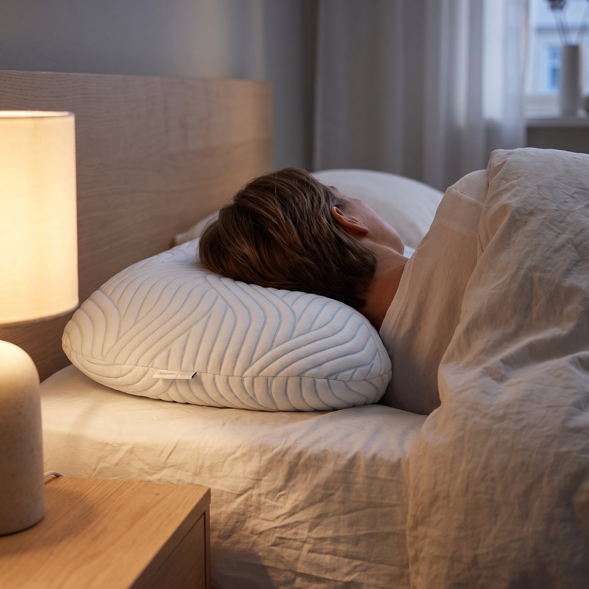 Person som sover på sidan med huvudet på en vit Tempur Sonata-kudde med ljusblått vågmönster, sedd bakifrån i ett sovrum med sänglampa