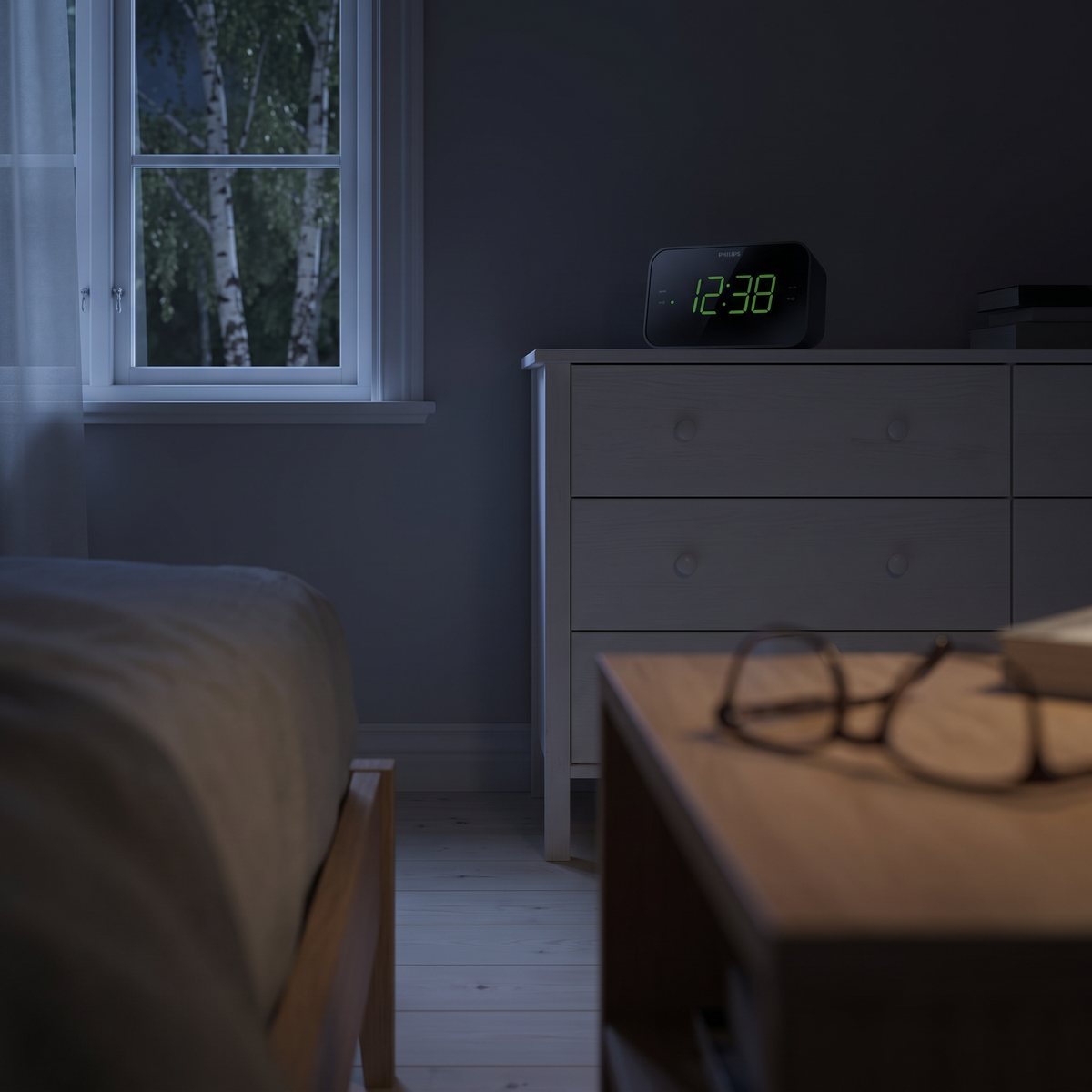 Bred svart klockradio Philips TAR3306 med stora gröna siffror på en vit byrå i ett mörkt sovrum, läsglasögon på nattduksbordet i förgrunden