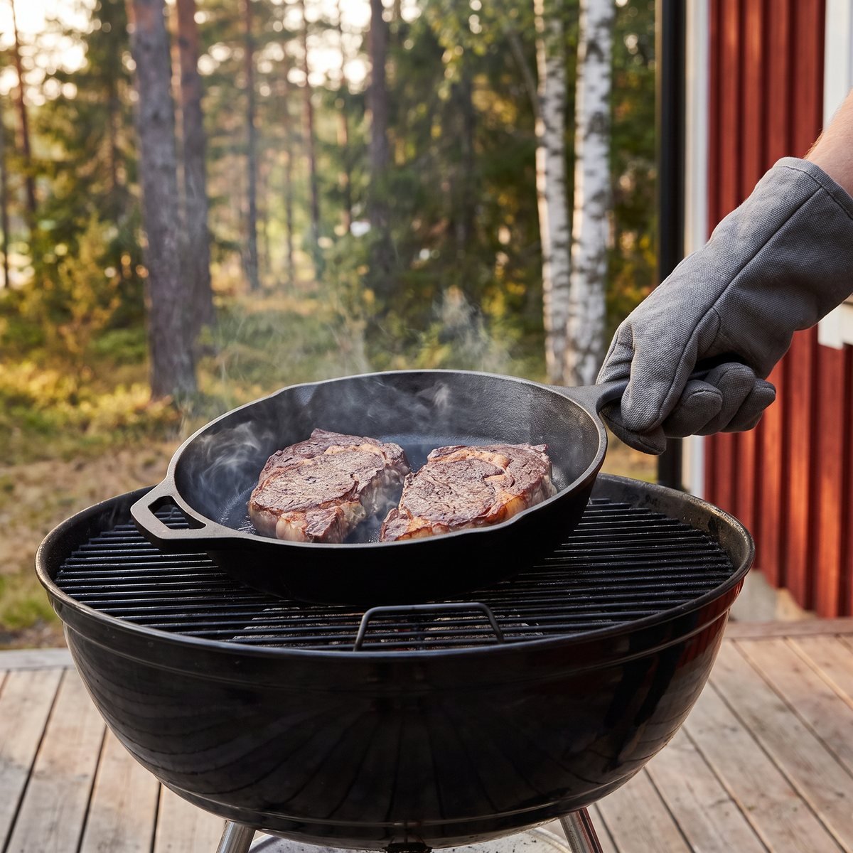 Lodge stekpanna i gjutjärn med två biffar på en kolgrill på en altan vid ett rött trähus med tallar och björkar