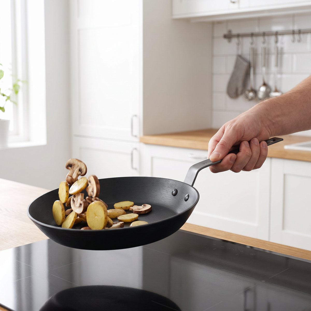 Ronneby Bruk Ultra Light Pro stekpanna med långt stålskaft som skakas med en hand ovanför en induktionshäll med svamp och potatis