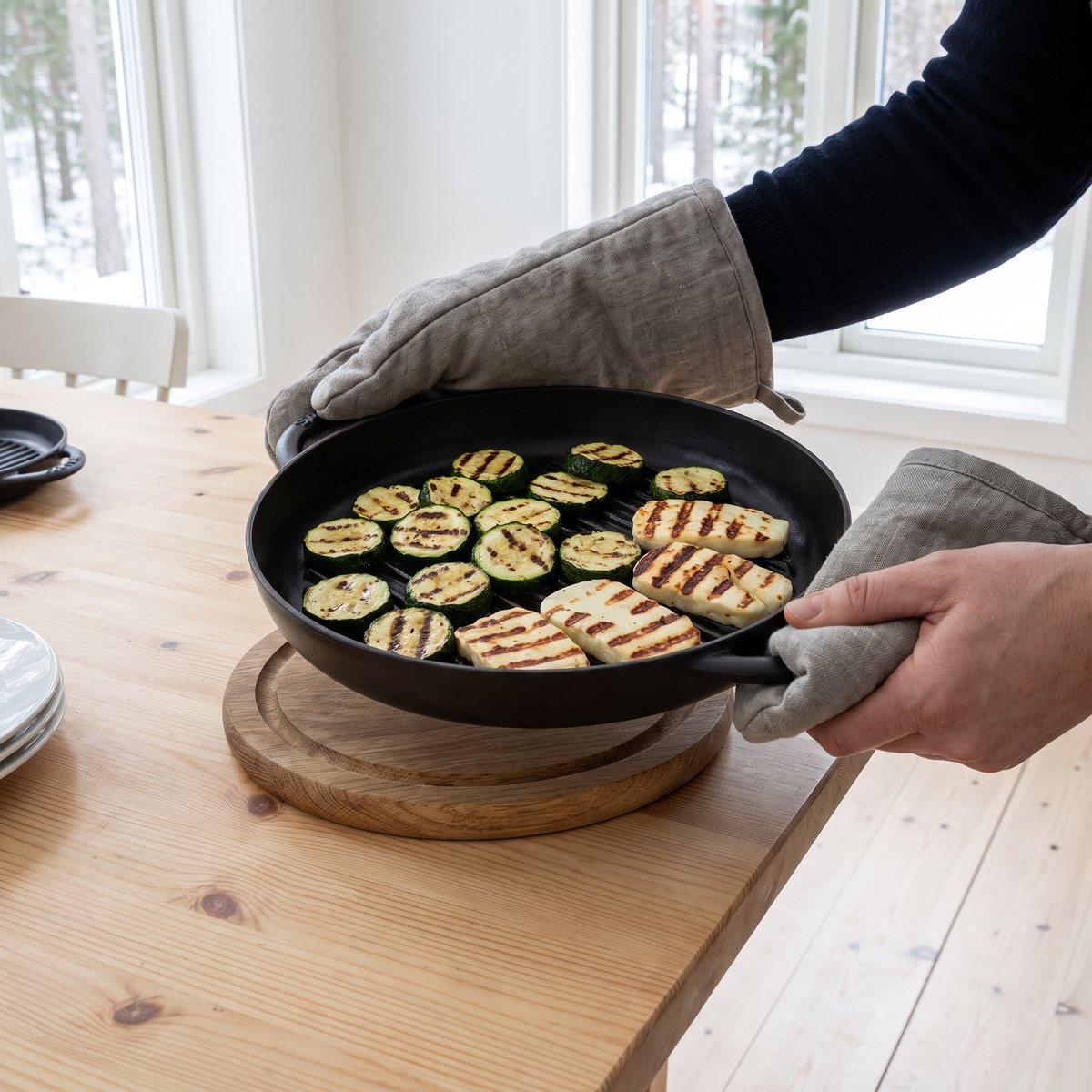 Staubs runda svarta grillpanna med två öron bärs med grytlappar till ett furubord, full med grillad zucchini och halloumi