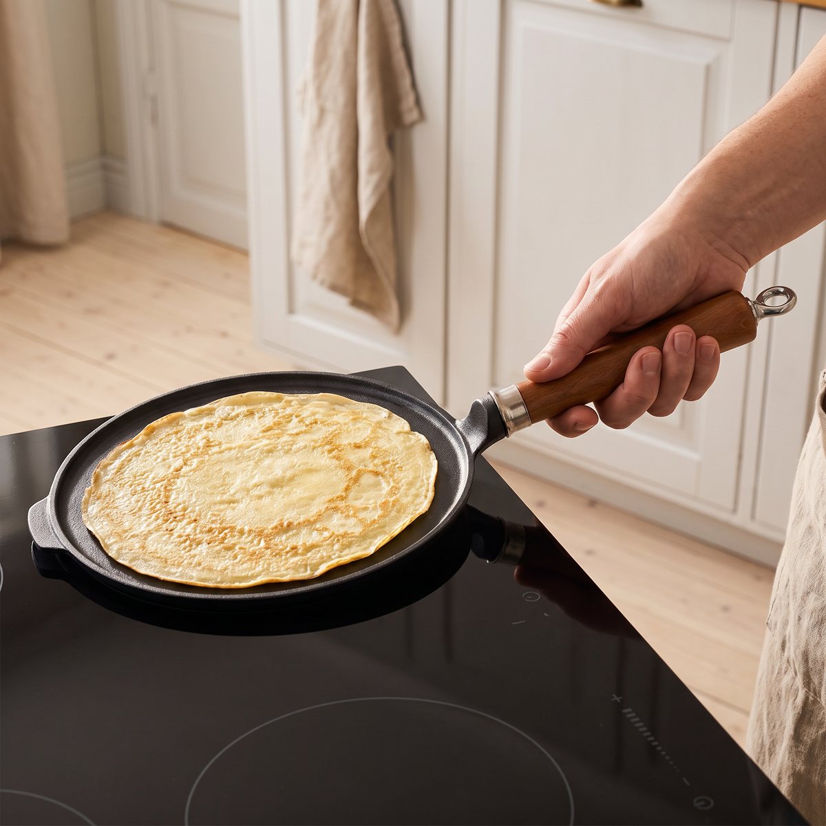 Ronneby Bruk Maestro pannkakspanna i gjutjärn hålls i träskaftet över induktionshällen i ett ljust svenskt kök