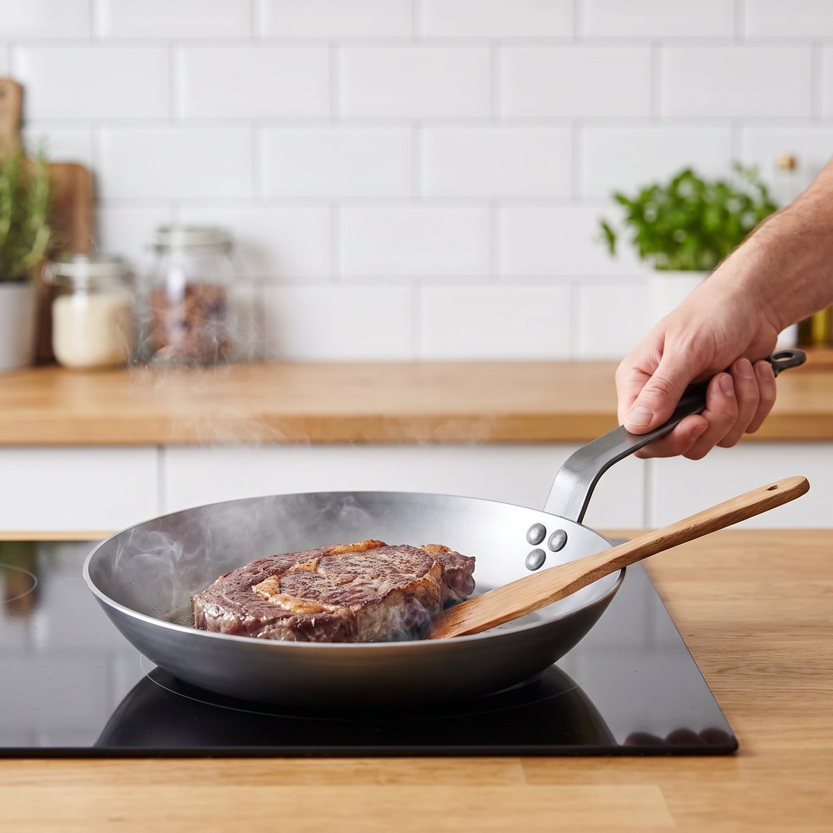 Blank kolstålspanna med långt stålhandtag där en entrecote steks på induktionshäll i ett svenskt kök med vita kakelplattor