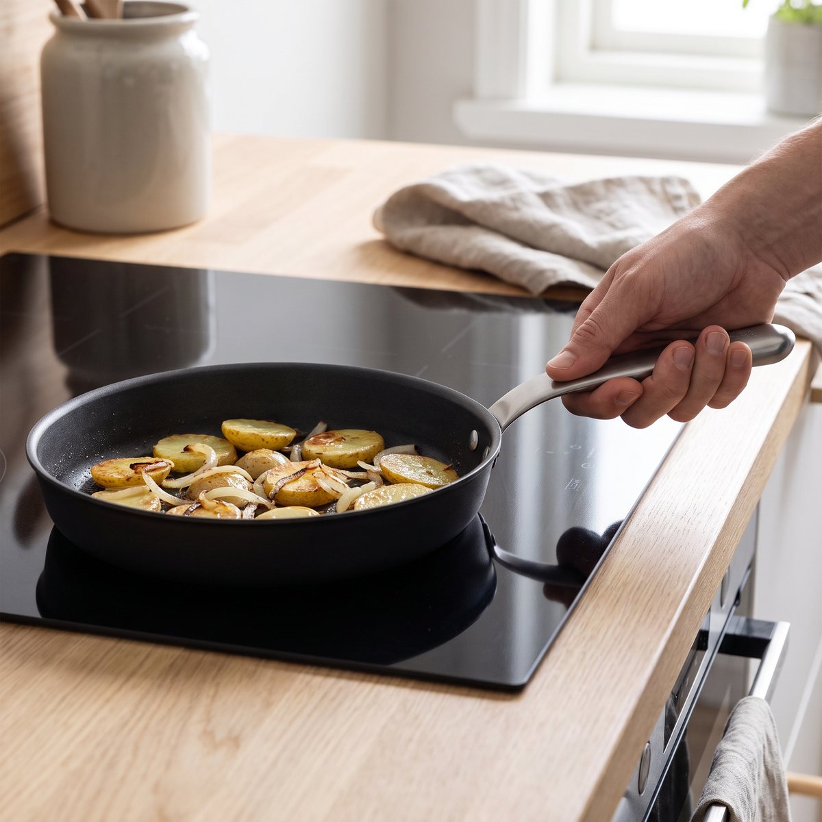 Mörk kolstålspanna med ergonomiskt stålhandtag där potatis och lök steks på induktionshäll i ett skandinaviskt kök