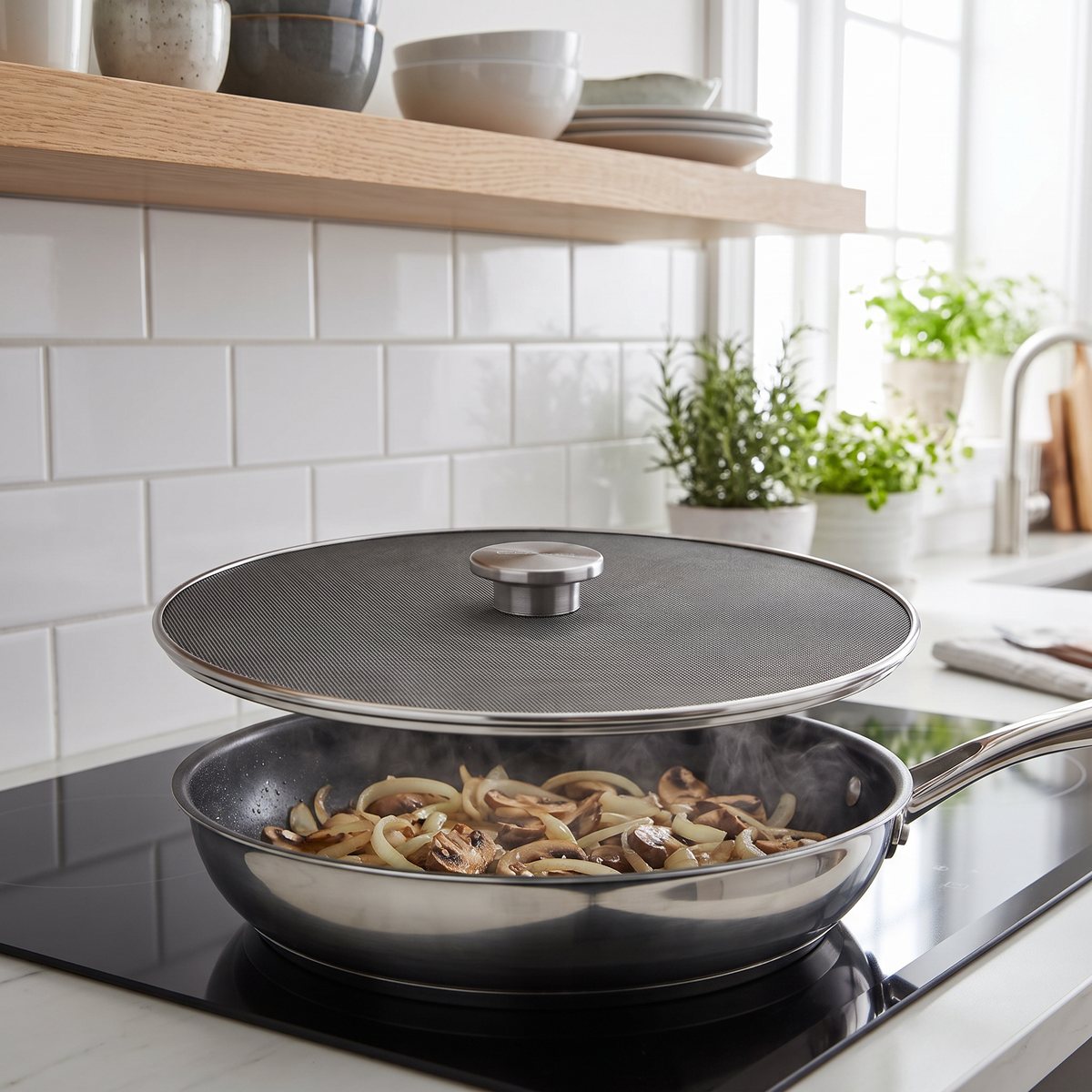 KitchenAid stänkskydd i mörkt nät med stålknopp över en stekpanna med lök och svamp på induktionshäll i ett svenskt kök