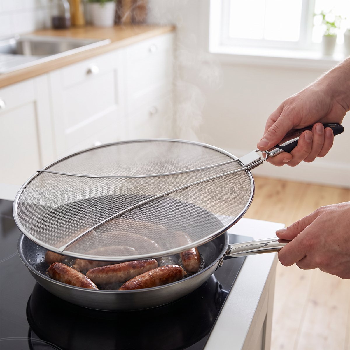 MasterClass stänkskydd med finmaskigt nät och handtag hålls över en stekpanna med korv på induktionshäll i ett svenskt kök