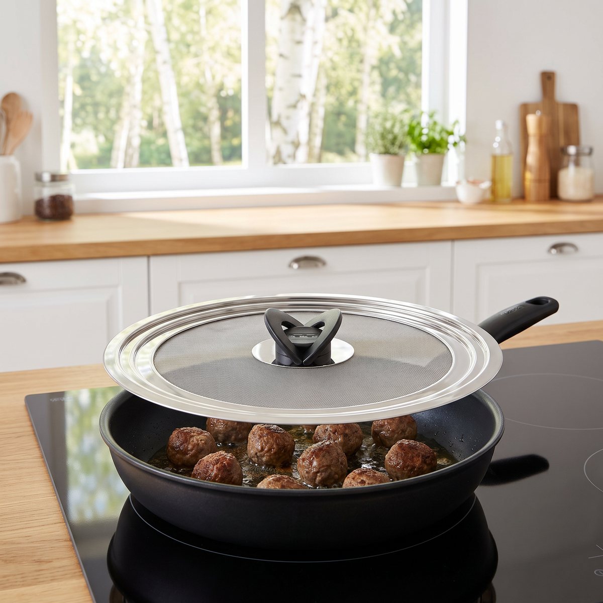 Tefal Ingenio stänkskyddslock av rostfritt stål med finmaskigt nät på en stekpanna med köttbullar på induktionshäll i ett svenskt kök