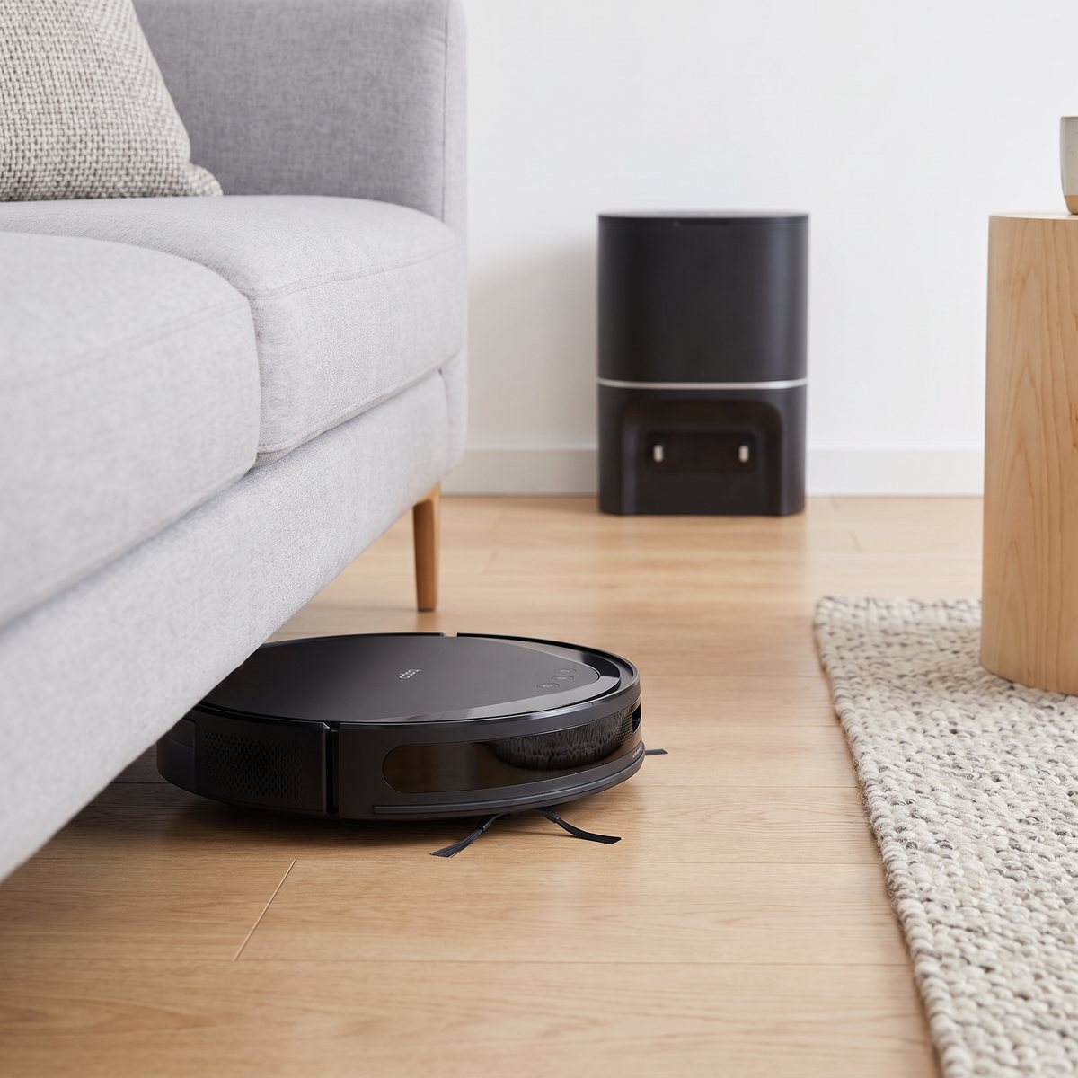Svart TP-Link Tapo RV20 Max Plus glider in under en ljusgrå soffa på ekgolv i ett svenskt vardagsrum, den svarta stationen vid väggen